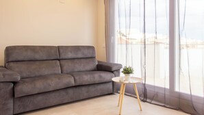 Ein Wohnzimmer mit grauem Sofa, einem kleinen runden Tisch mit Pflanze und einem großen Fenster mit transparenten Vorhängen.