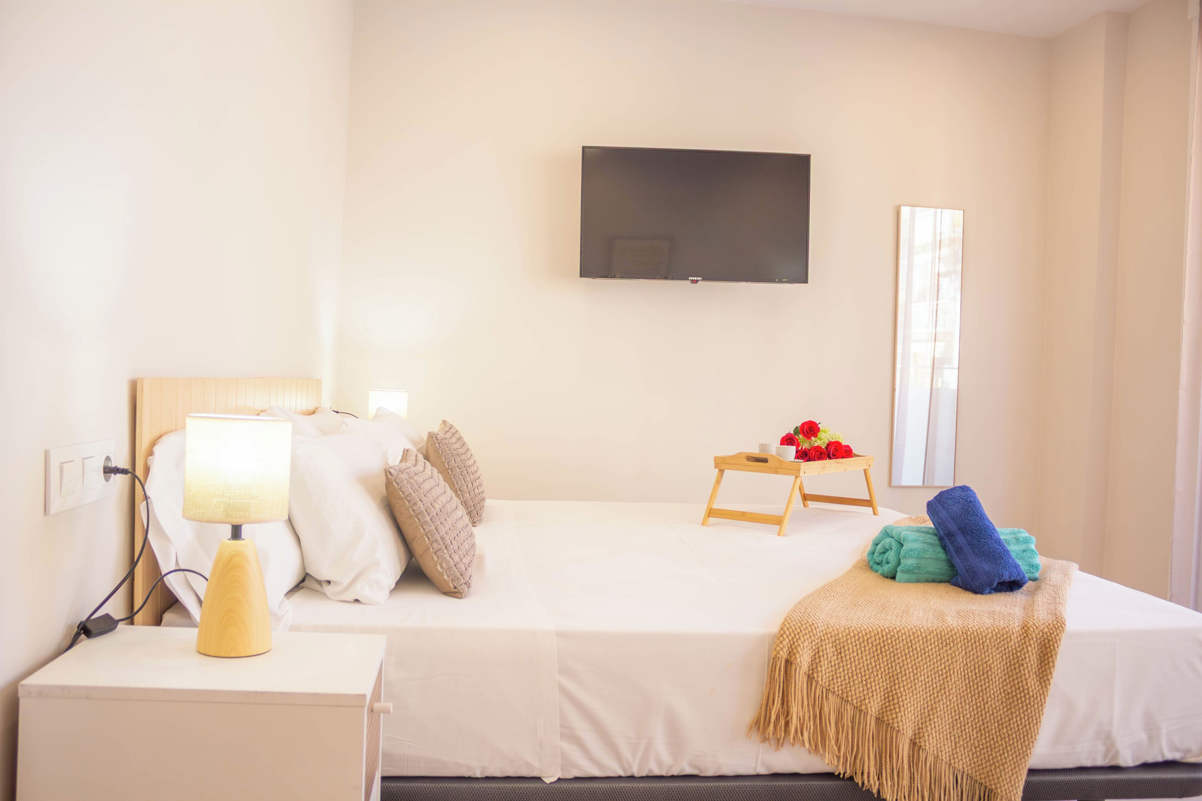 Habitación de hotel con cama, mesita de noche con lámpara, televisor montado en la pared, pequeña mesa de madera con flores y ventana con cortinas.