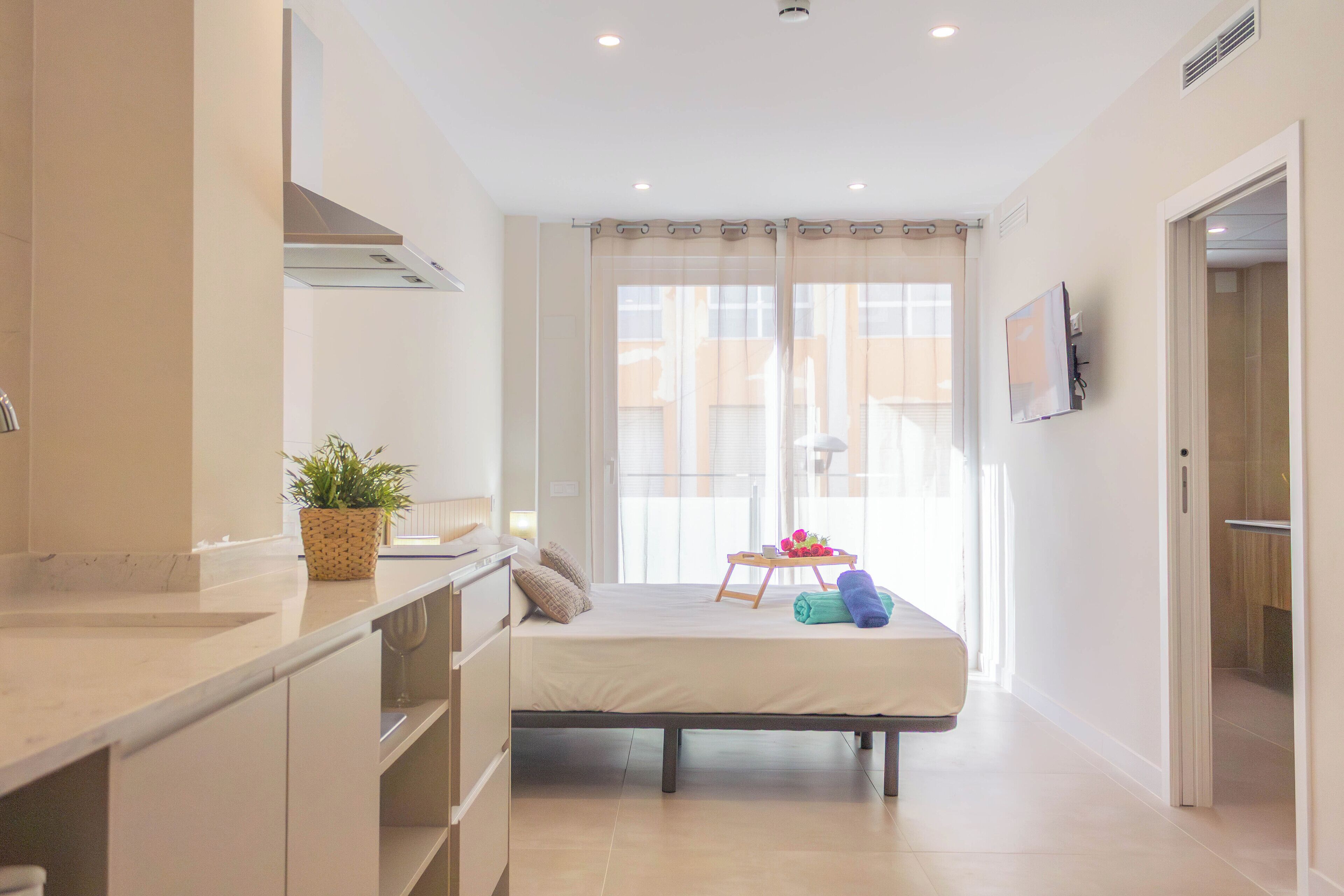 Habitación de hotel moderna con kitchenette, una cama con mesita y toallas, y un ventanal grande con cortinas.