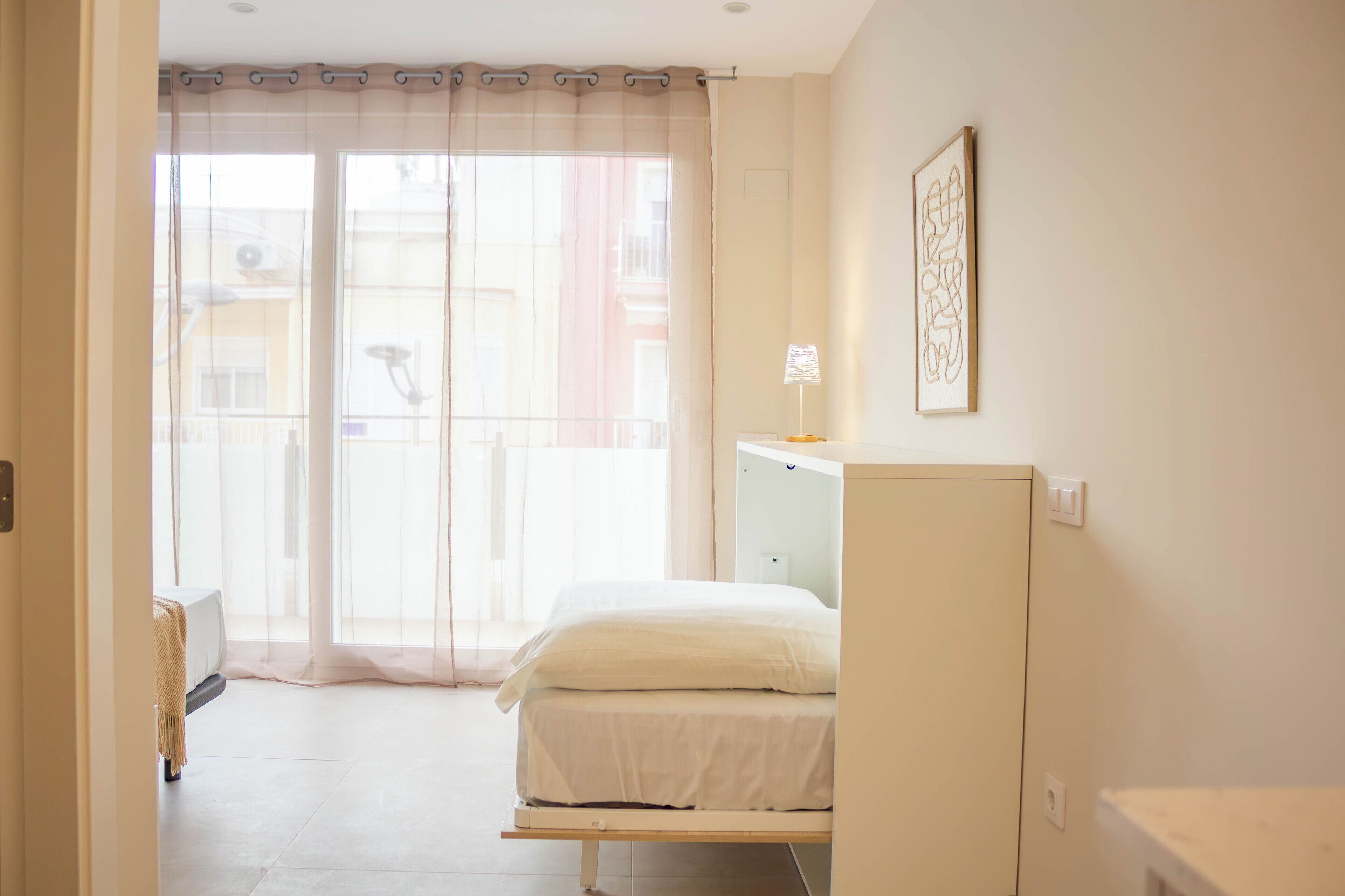 Une chambre à coucher avec un lit, une table de chevet, une lampe et une fenêtre avec des rideaux légers.