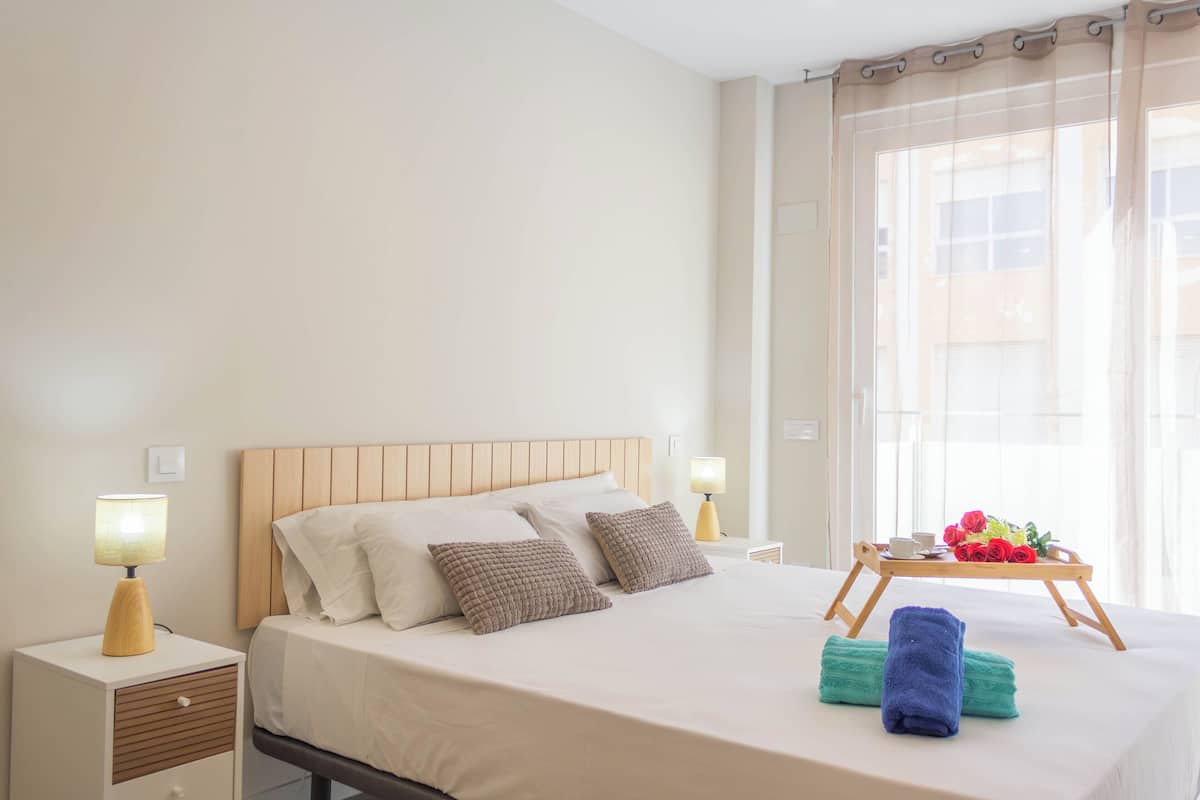 Una cama bien hecha con almohadas, una mesita de noche con lámpara y una bandeja con flores y una taza en la mesita de noche.