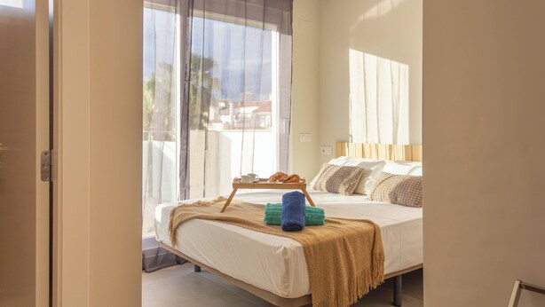 Ein Hotelzimmer mit Bett, Vorhängen am Fenster, einem Nachttisch mit Tablett und Blick ins Freie.