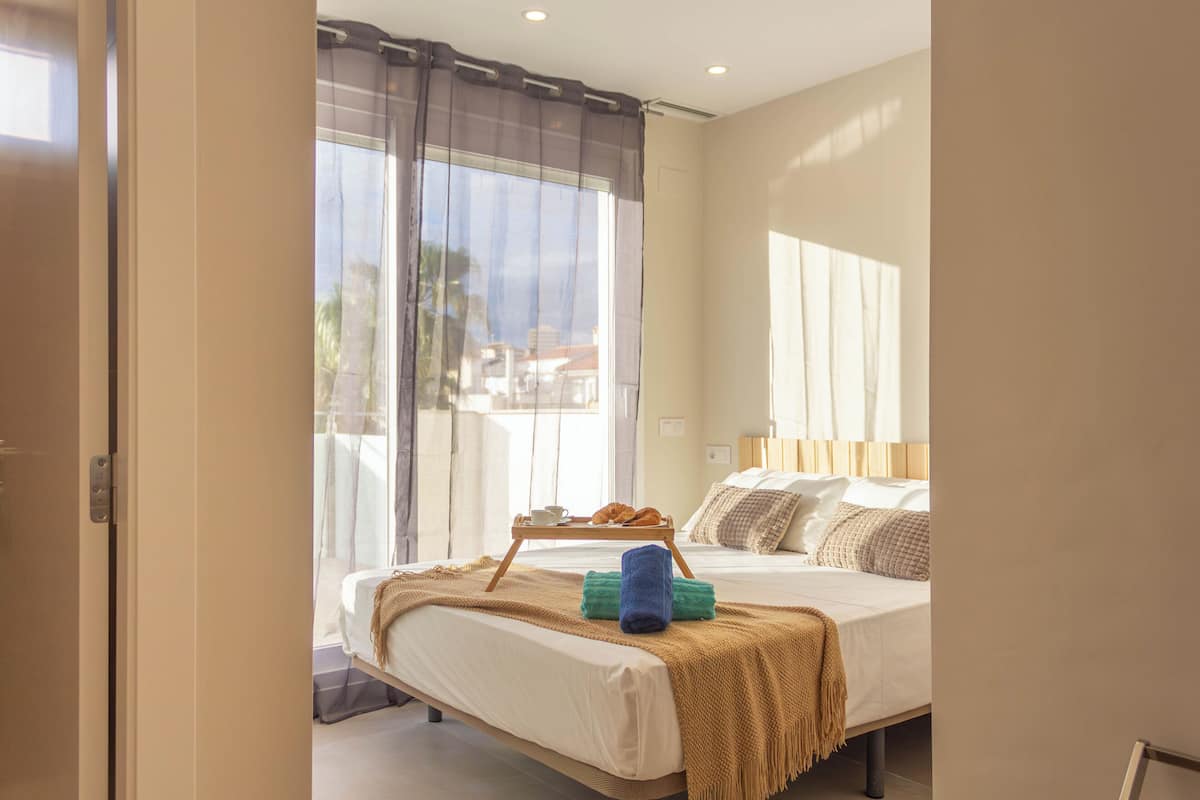 Habitación de hotel con cama, ventana con cortinas, mesita de noche con bandeja y vistas al exterior.