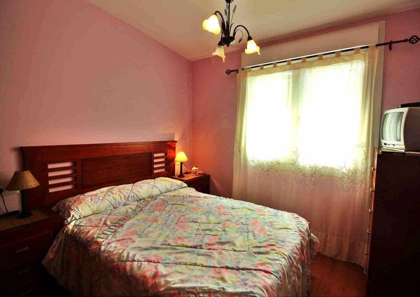 Un dormitorio con una cama, una mesita de noche con lámpara, un televisor sobre un mueble y una ventana con cortinas.