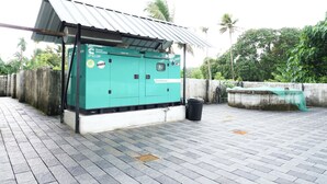A power generation unit labeled "SilentPower" with a metal roof, situated on a paved area with a concrete wall and a palm tree in the background. - Charming 2-bedroom AC apartment Close to Rajagiri Hospital (KL)