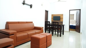 A modern living room with a brown leather sofa, a wooden dining table with chairs, and a ceiling fan. - Charming 2-bedroom AC apartment Close to Rajagiri Hospital (KL)