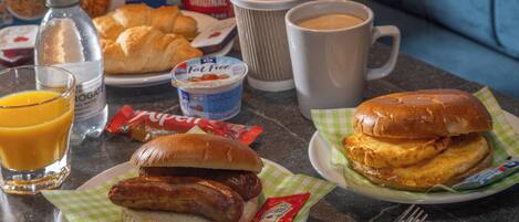 Una mesa de desayuno con croissants, salchichas, un sándwich, jugo, café y cajas de cereal.