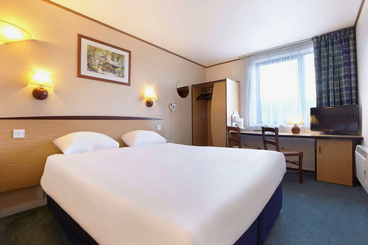 A hotel room with a large bed, a desk with a chair, a television, and a window with curtains.