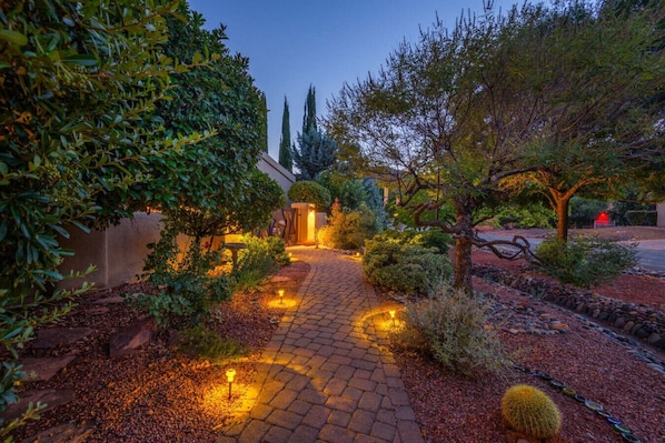 A paved garden pathway leading to a building entrance, surrounded by various shrubs and trees.