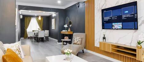 A modern living room with a flat-screen TV mounted on a marble wall, a sofa, a coffee table, and a dining area in the background.