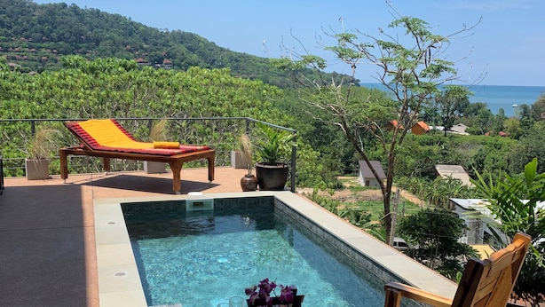 A pool area with a wooden lounge chair, a table with a vase of flowers, and a view of a lush landscape and ocean. - Boho-Luxe Pool Villa with Simply Amazing Views by Setting Sun Boutique Homes (ตำบล เกาะลันตาใหญ่)