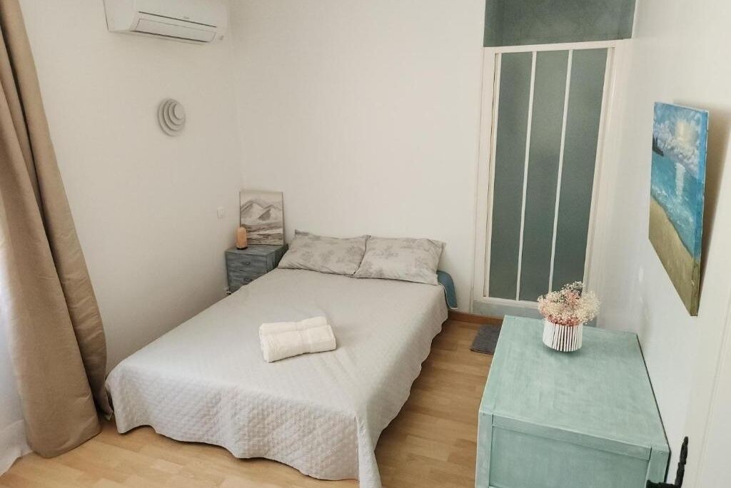 Ein kleines, aufgeräumtes Schlafzimmer mit einem Bett, einem Nachttisch, einem Fenster mit Jalousien und einem kleinen Tisch mit einer Vase.