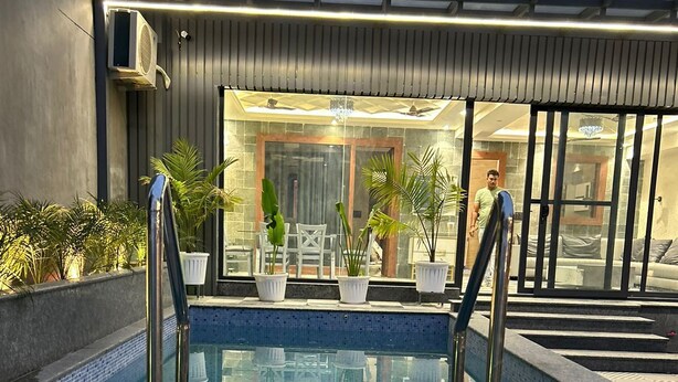 An outdoor swimming pool with a glass barrier, surrounded by potted plants and a modern building with a balcony. - Ganga Euphoria Villa (Rishikesh)