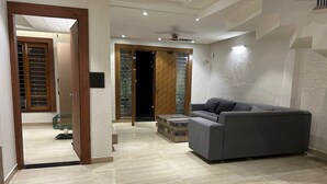 A modern living room with a grey sofa, a glass coffee table, and a wooden door leading to another room. - Ganga Euphoria Villa (Rishikesh)
