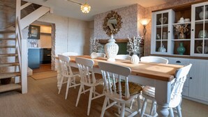 Un coin repas avec une table en bois et des chaises blanches, un escalier et une cuisine visible au fond.