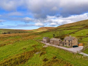 A stone farmhouse with a stone wall and a small garden, surrounded by rolling hills and sheep grazing.