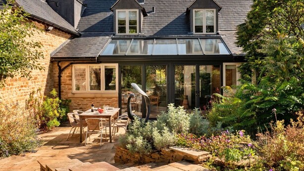 Ein Haus mit Wintergarten, Steinmauern und einem Garten mit einer Patio-Bestuhlung.