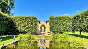 Ein friedlicher Garten mit einem zentralen Brunnen, umgeben von ordentlich geschnittenen Hecken und blauem Himmel.