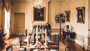 Un comedor formal con una larga mesa dispuesta para una comida, rodeada de sillas, un candelabro y retratos enmarcados en las paredes.
