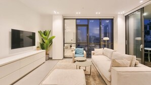 A modern living room with a flat-screen TV, a large potted plant, a sofa, a coffee table, and a view of the city through large windows. - Hayarkon Bronze in Tel Aviv-yafo (Tel Aviv)
