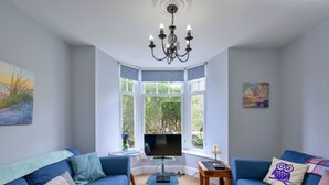 Een woonkamer met twee blauwe banken, een televisie, een kroonluchter en grote ramen met uitzicht op groen.