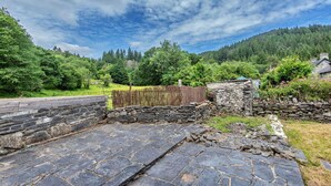 Un chemin de pierres mène à un bâtiment en pierre, bordé d’une clôture en bois et entouré d’une végétation luxuriante.