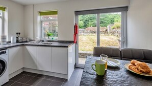 Une cuisine moderne équipée d’un lave-linge intégré, un coin repas avec une table pour deux personnes et une vue sur un jardin grâce à de grandes fenêtres.