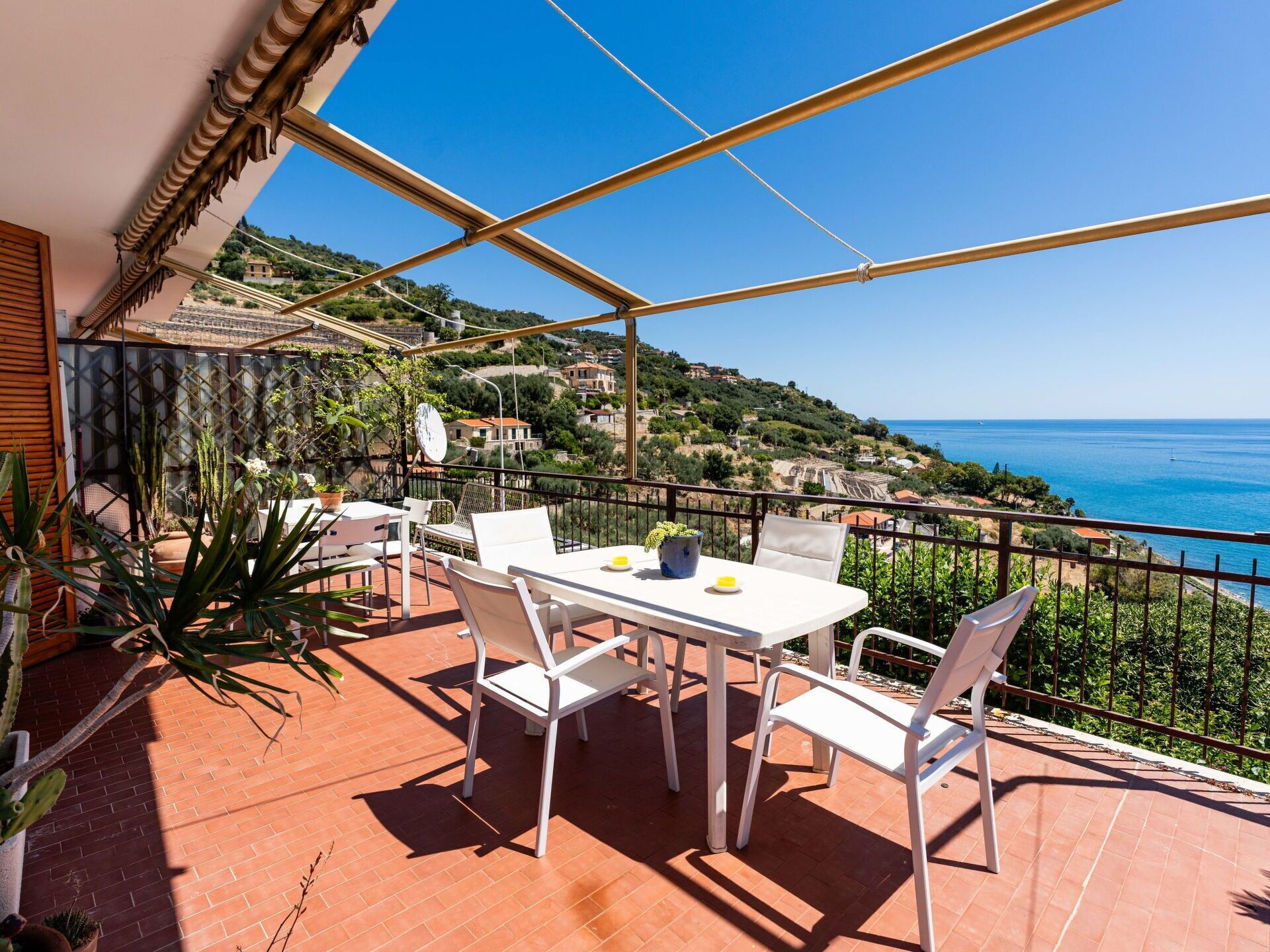 Eine Terrasse mit einem weißen Tisch und Stühlen, Kübelpflanzen und Blick auf das Meer und die Hügel.