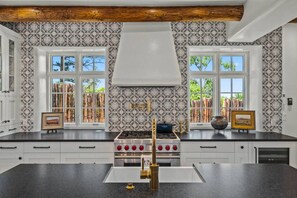 A kitchen with a large island, double sinks, and a gas stove. There are two windows with a view of trees and a wooden ceiling.