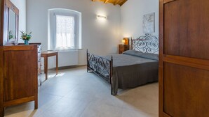 A bedroom with a wooden ceiling, a bed with a metal headboard, a bedside table, a dresser, a window with a curtain, and a wall-mounted light. - Beautiful apartment for 7 people with A/C, WIFI and pets allowed (Donoratico)