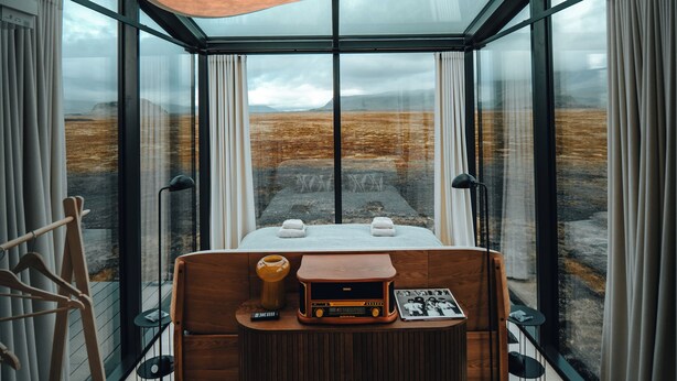 A modern bedroom with a large window, a wooden bed, a small table with a radio, and a hanging light fixture. - Glass-roof lodge with private spa & sauna near Seljalandsfoss, pure tranquility (Hvolsvöllur)