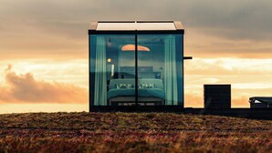 A modern, cube-shaped glass structure with a bed and a desk, set against a sunset sky. - Glass-roof lodge with private spa & sauna near Seljalandsfoss, pure tranquility (Hvolsvöllur)