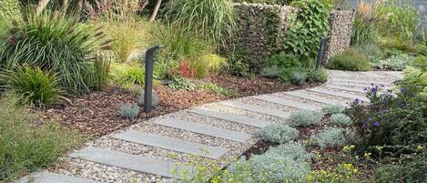 A garden pathway with a stone border, surrounded by various plants and a stone wall.