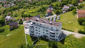 A multi-story building with balconies, surrounded by green fields and houses.