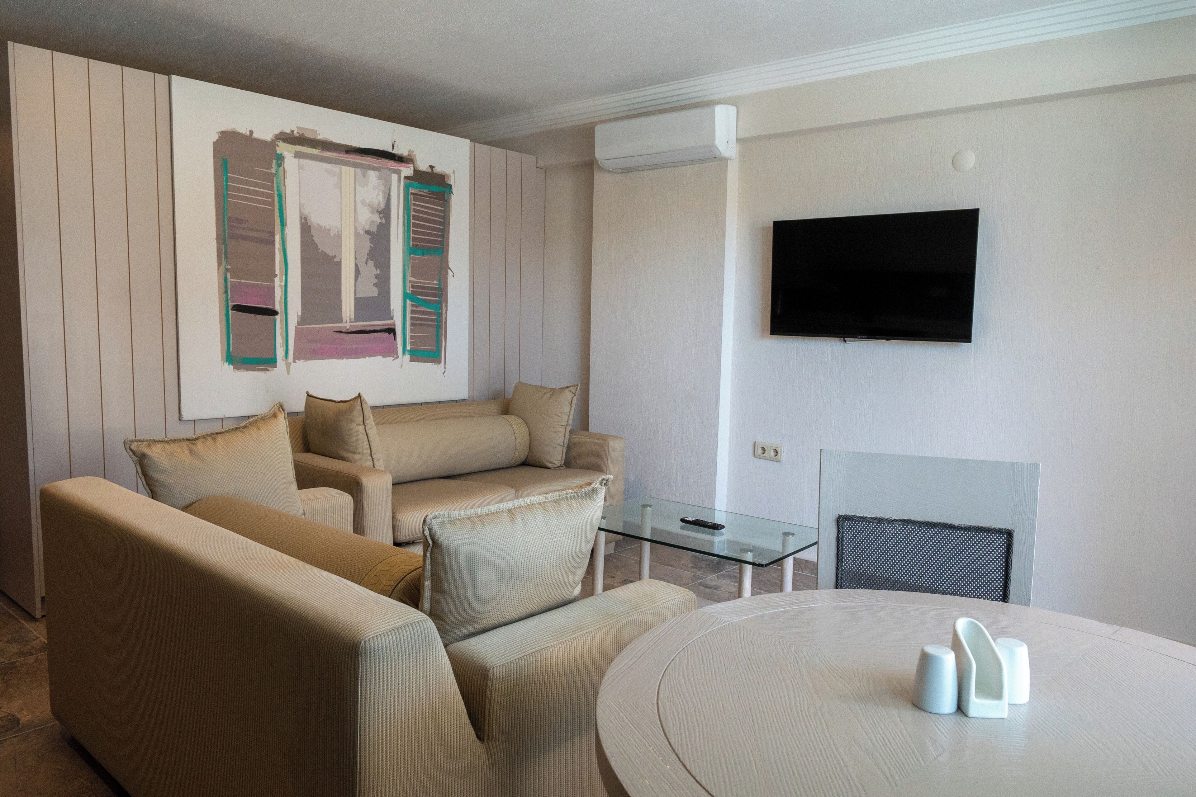 Een woonkamer met een beige bank, een glazen salontafel en een flatscreen televisie aan de muur.