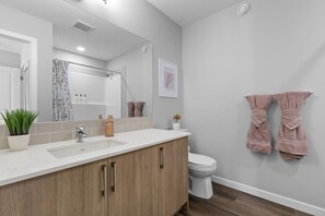 Une salle de bain moderne avec un grand miroir, un lavabo, des toilettes et une douche. Des serviettes sont accrochées au mur et une plante en pot est posée sur le comptoir.