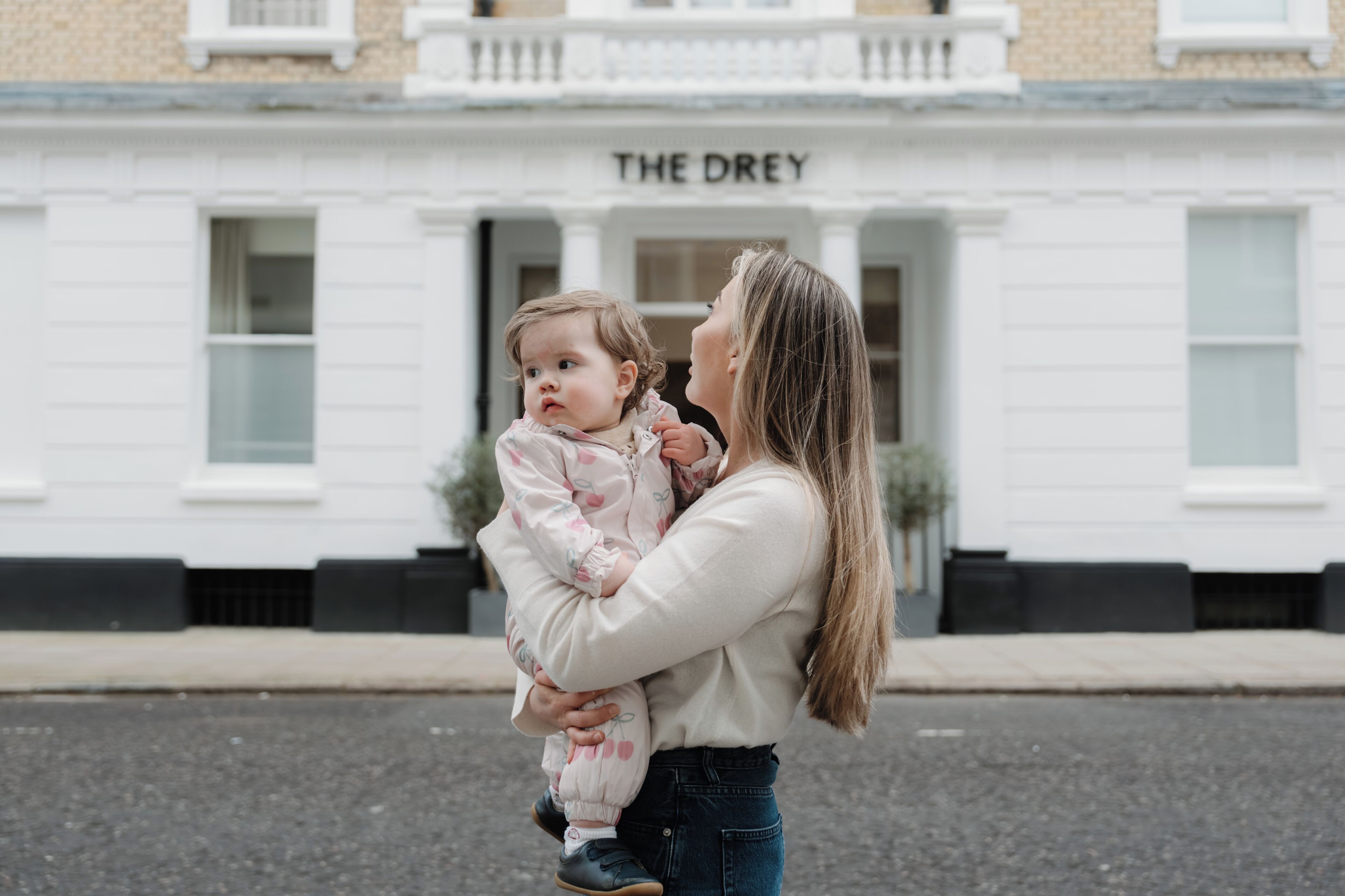 A woman holding a child in front of a building with 'THE DREY' sign.