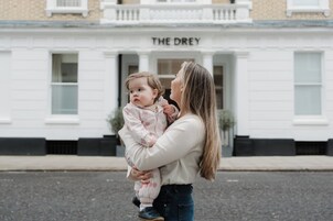 Een vrouw die een kind vasthoudt voor een gebouw met het bord 'THE DREY'.