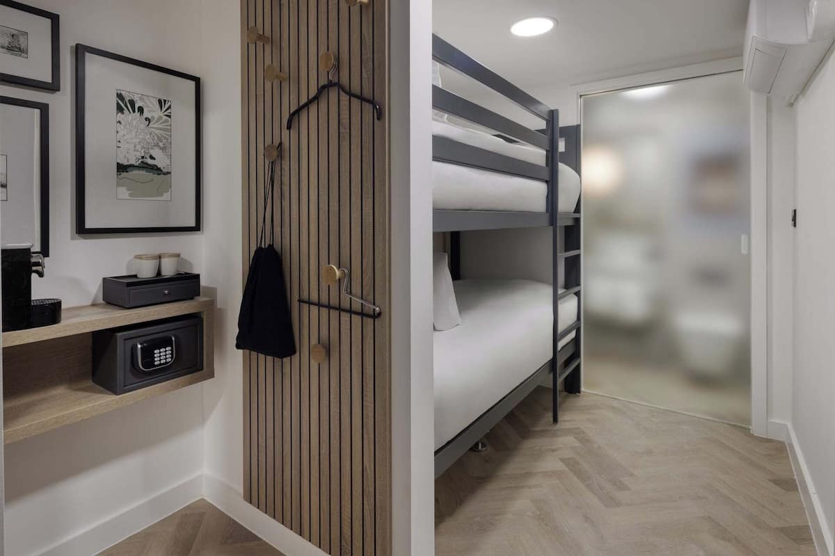 A compact hotel room with bunk beds, a wooden panel wall, a small desk with a coffee machine, and framed artwork.