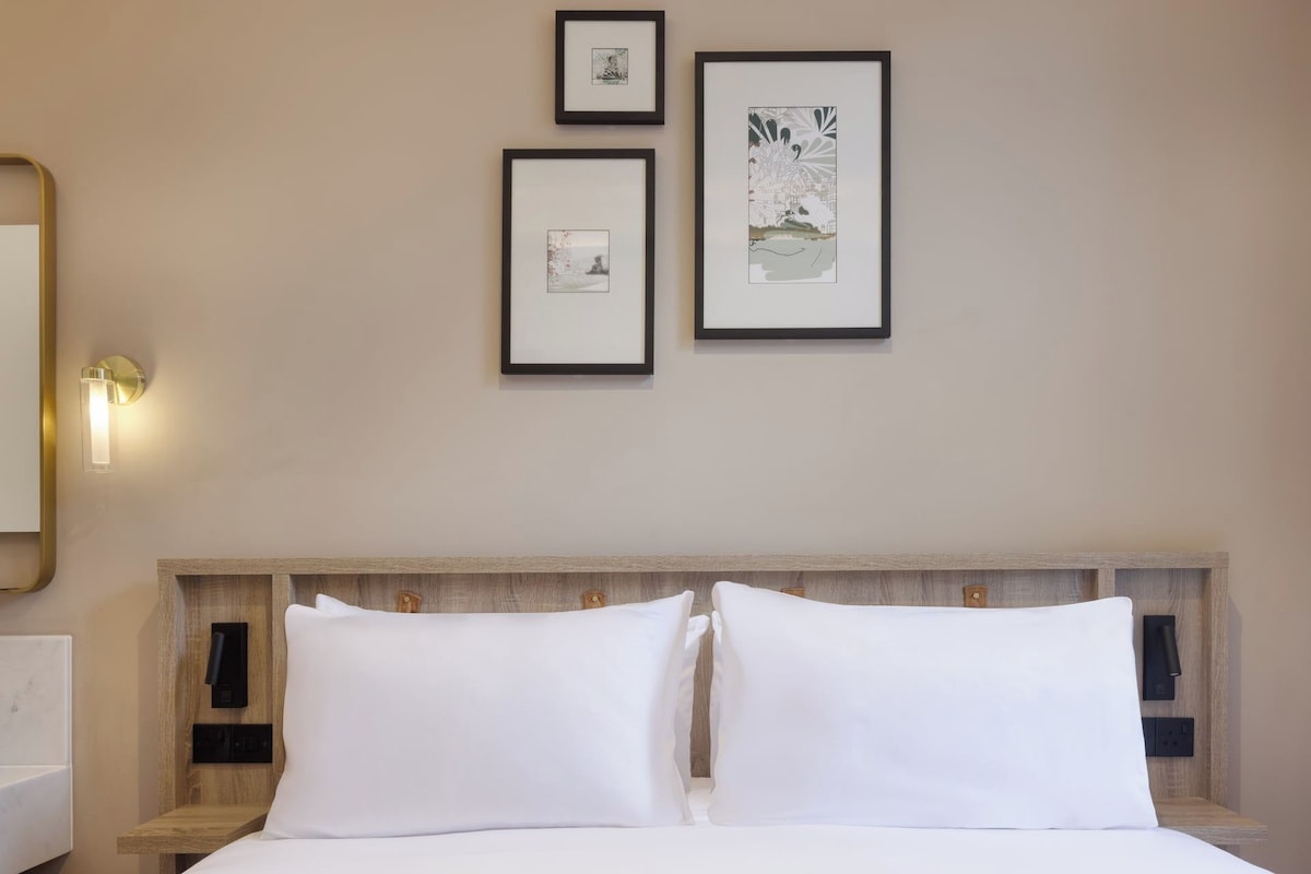 A hotel room with a bed, two pillows, a nightstand, a wall-mounted light, and three framed pictures on the wall.