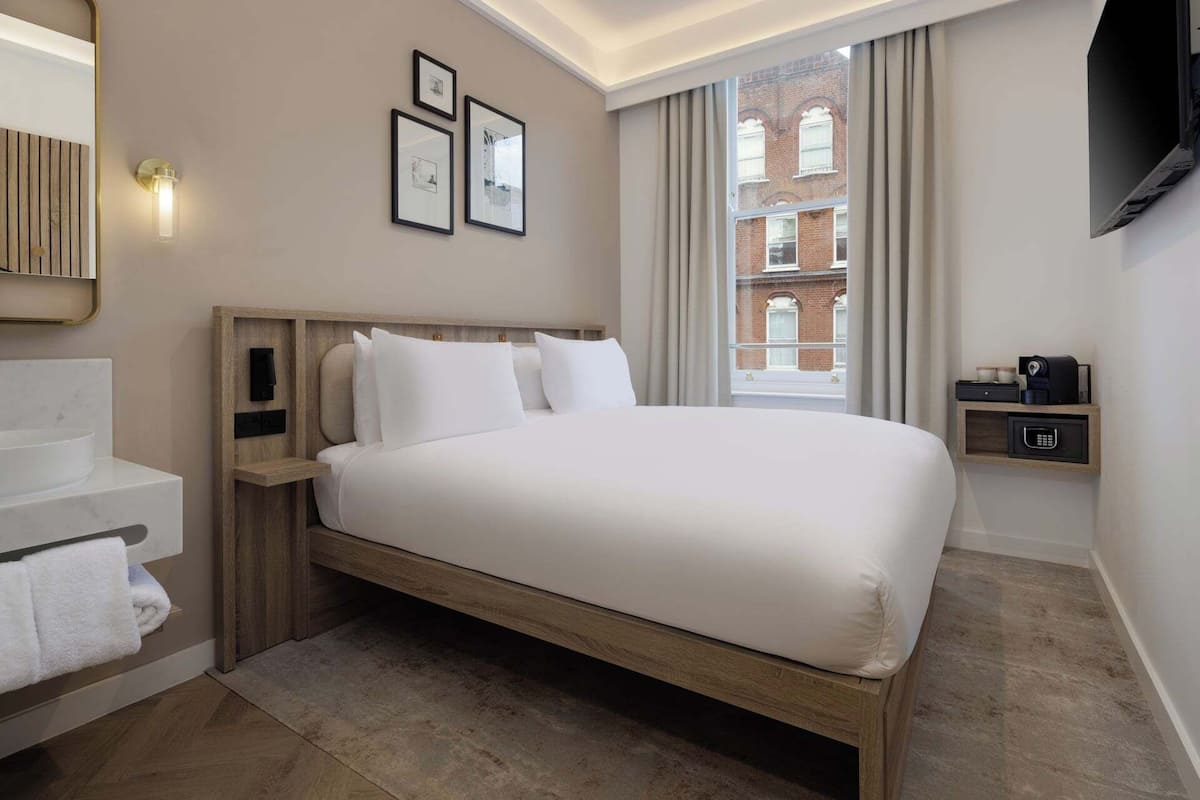 A hotel room with a bed, a vanity with a mirror, a small shelf with a coffee maker, and a window with curtains.