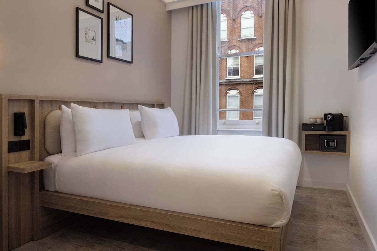 A hotel room with a bed, a bedside table, a TV, and a window with a view of a brick building.