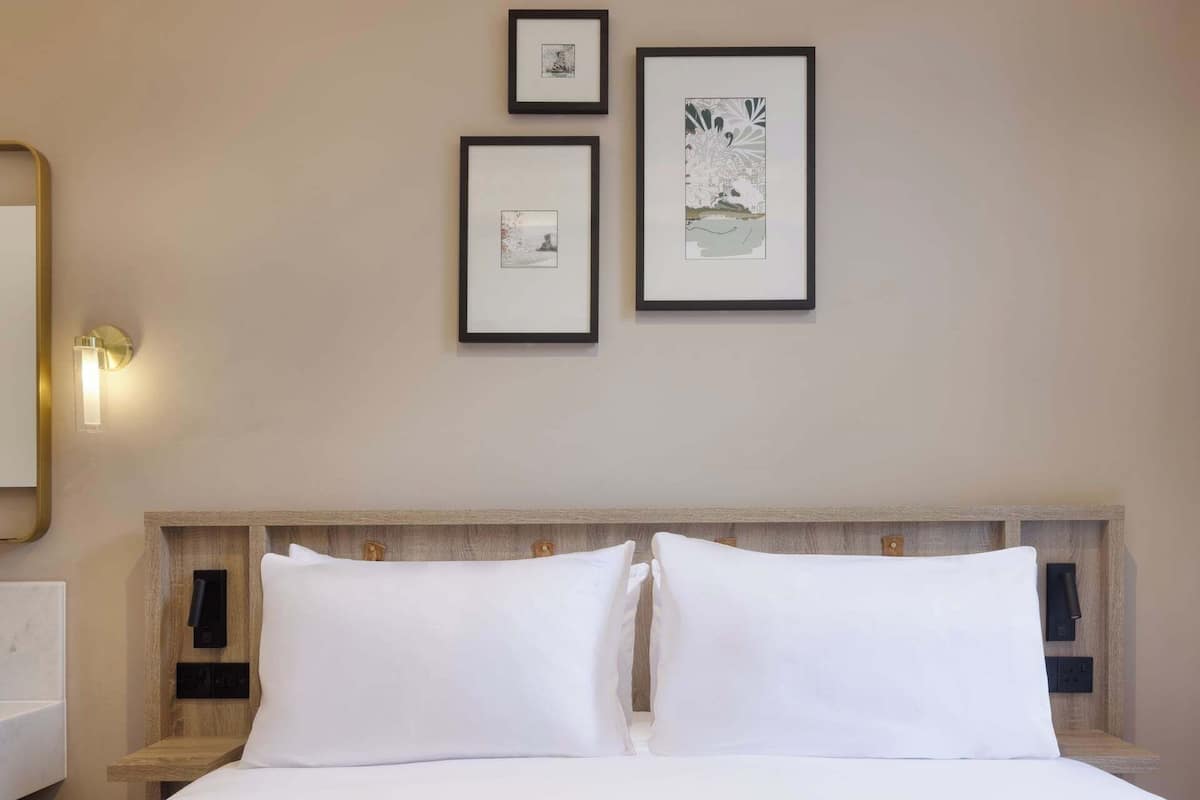 A hotel room with a bed, two pillows, a nightstand, a wall-mounted light, and three framed pictures on the wall.