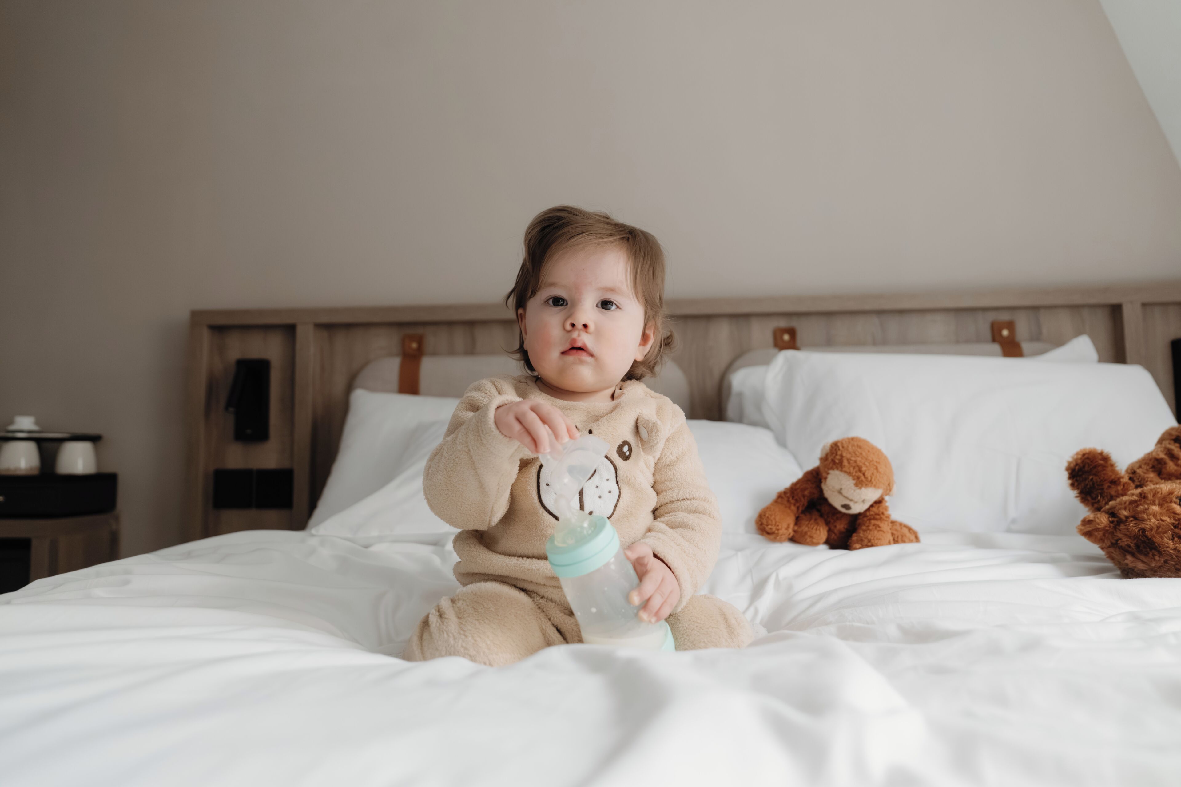 A child sitting on a bed with a teddy bear and a bottle.