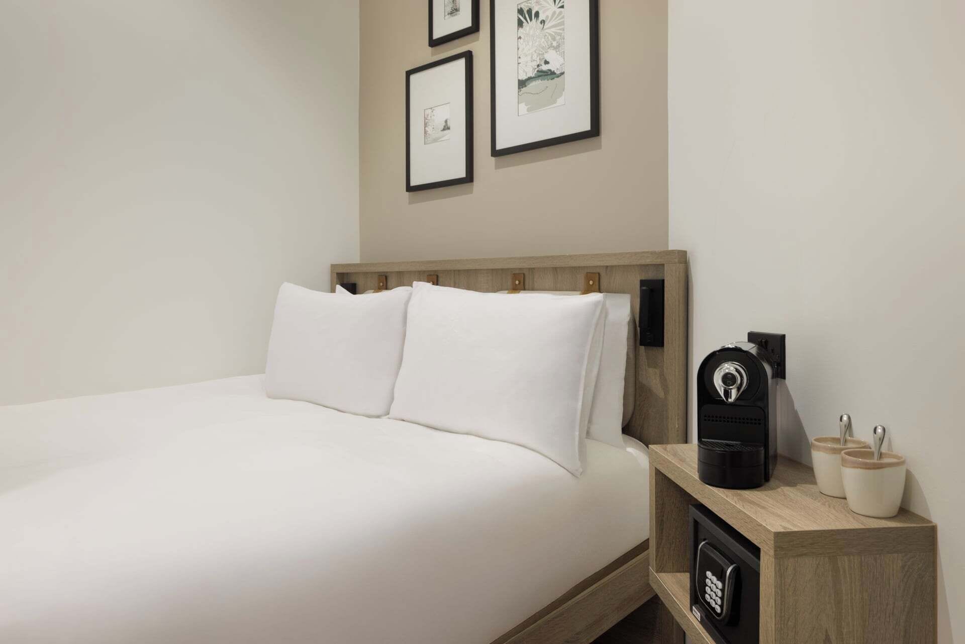 A neatly made bed with white linens, a wooden headboard, and a bedside table with a coffee maker and a cup.
