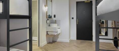 Una habitación de hotel moderna con literas, un baño con lavamanos y espejo, y una puerta que da a otra habitación.
