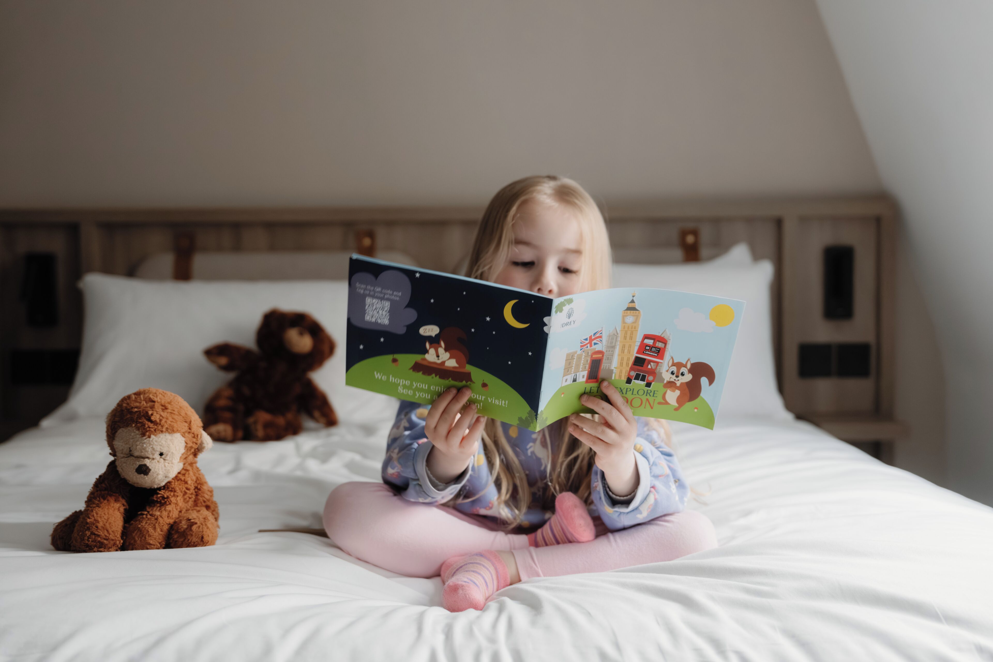 A child reading a book on a bed with a teddy bear beside her.