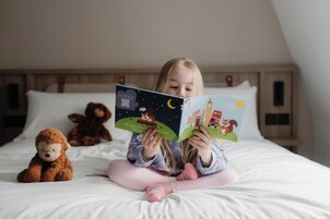 Ein Kind liest ein Buch im Bett, neben ihr ein Teddybär.