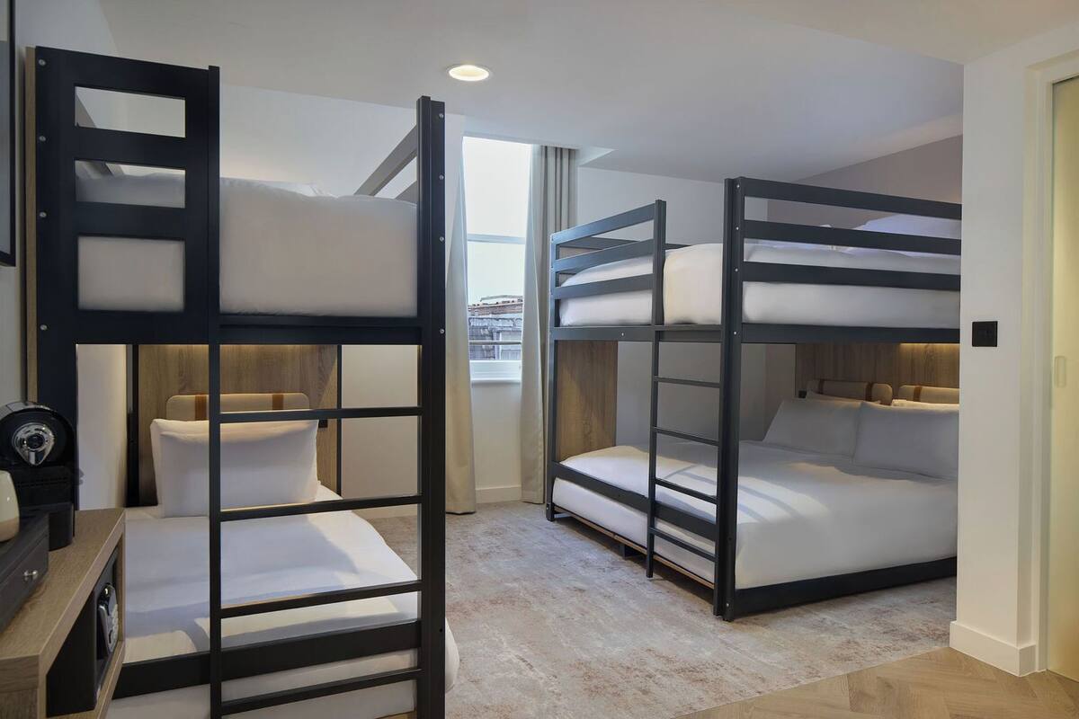 A compact hotel room with bunk beds, a small seating area, and a view through a window.
