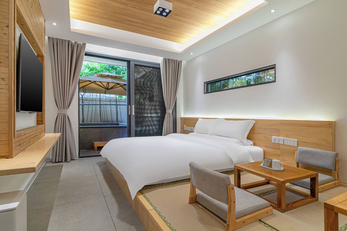A modern hotel room with a large bed, a wooden desk, a chair, a TV, and a window with a view of greenery.